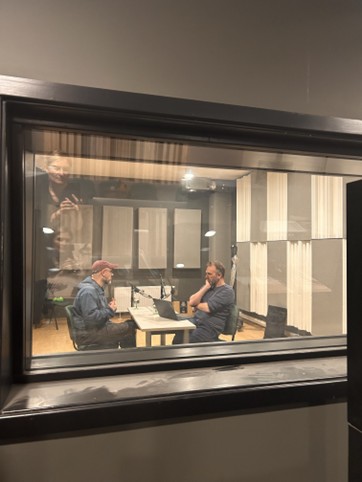 Two people sit across from each other at a table in a soundproof recording studio, seen through a window. They appear to be having a conversation or recording a podcast, with microphones and a laptop on the table.