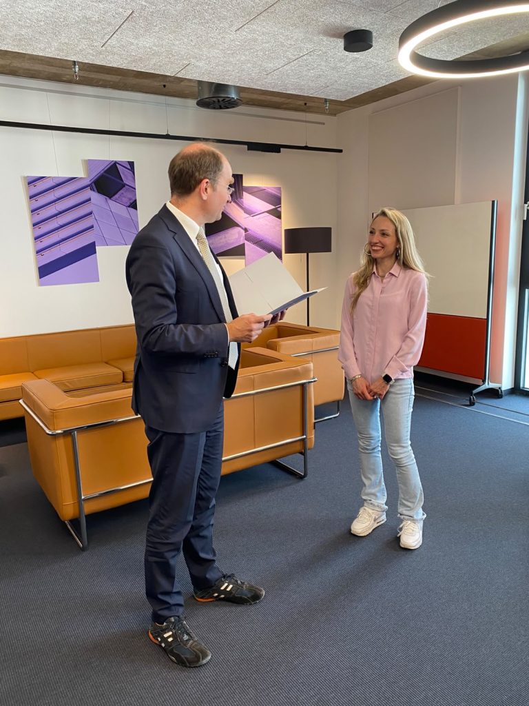 A man in a suit holding a folder talks to a smiling woman in a pink shirt and jeans in an office lounge.