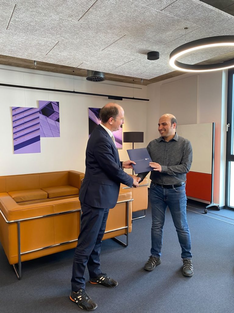 A man in a suit shakes hands with a man in a grey shirt and jeans while exchanging a blue folder.