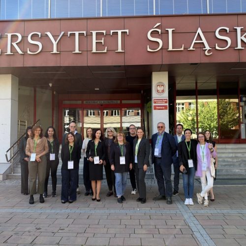 A group of people pose in front of the entrance to a building with the sign UNIWERSYTET ŚLĄSKI above them. Most wear conference badges and lanyards, and the weather appears sunny.
