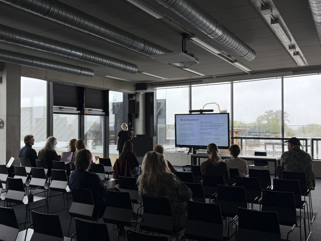 A presenter stands before a screen displaying a presentation in front of a seated audience in a conference room.