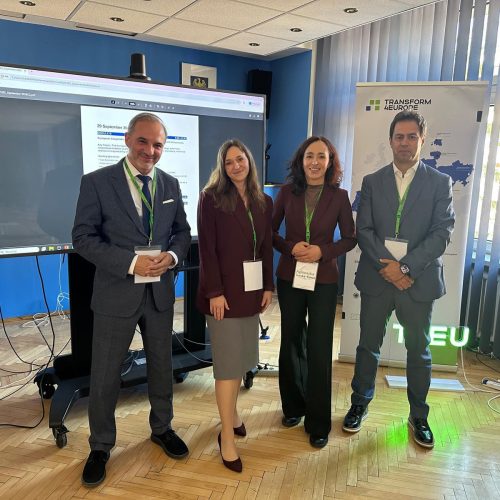 Four people in business attire stand and smile for a photo in a conference room. A digital screen and a Transform4Europe banner are visible in the background. All are wearing conference badges and green lanyards.