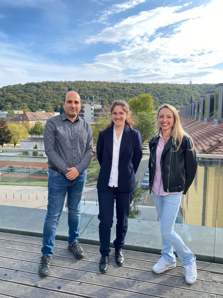 Three people standing side-by-side on an outdoor deck overlooking a town and a wooded hill.