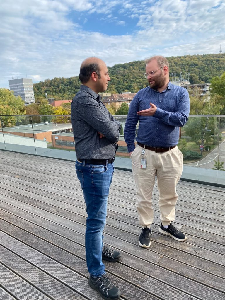 Two men stand on an outdoor wooden deck talking to each other with a cityscape and trees in the background.