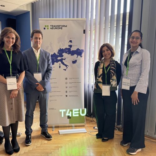 Four people stand indoors, smiling, beside a Transform4Europe banner showing a map of Europe, all wearing conference badges and green lanyards, with blinds and sunlight in the background.