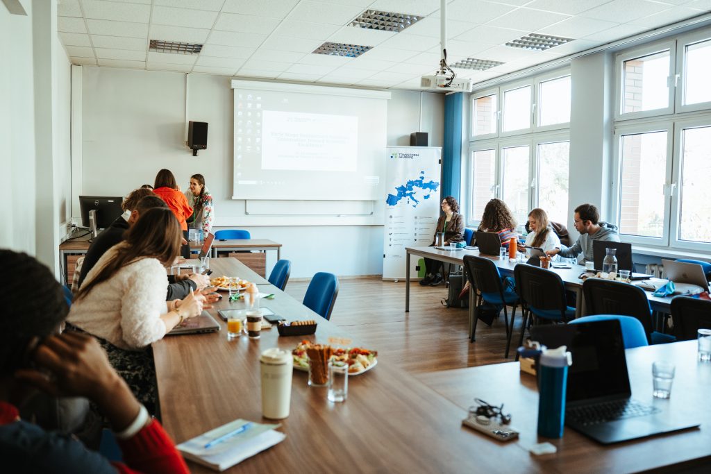 People attend a conference in a bright meeting room with tables, laptops, snacks, and a projector screen displaying presentation text.