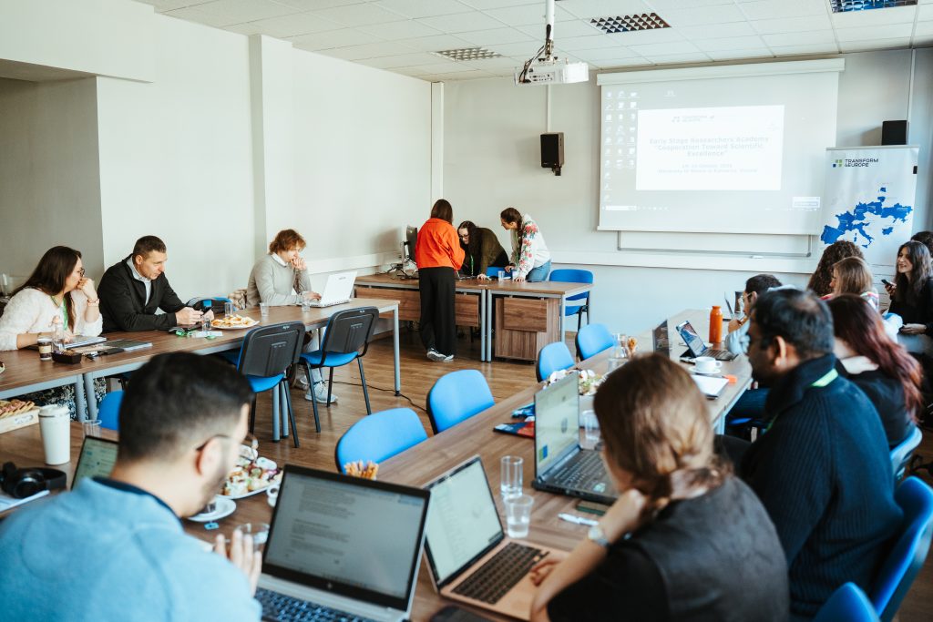 People sit at tables in a bright room working on laptops during a workshop, with a projection screen displaying Early Stage Researchers Academy at the front.