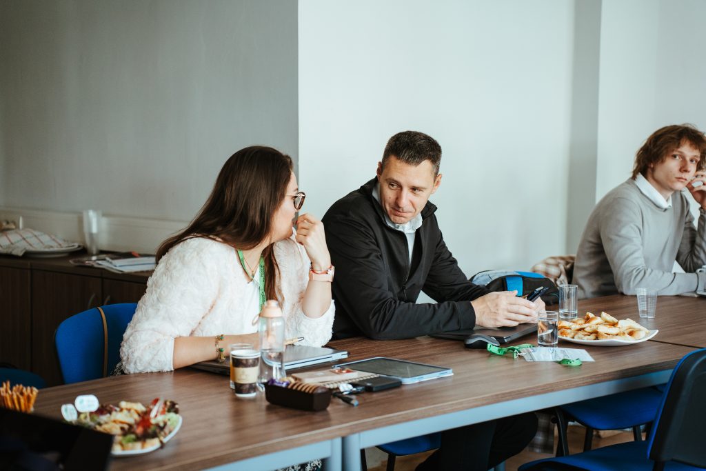 A woman and man sit at a table in a meeting room talking to each other, with another man sitting nearby and plates of food on the table.