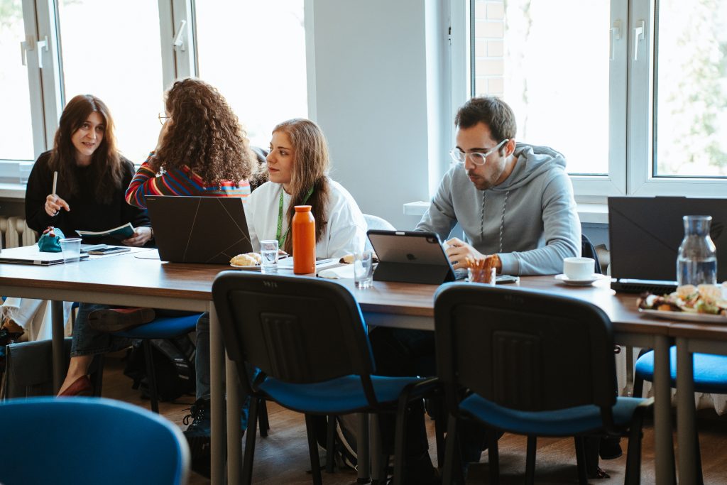 Four people sitting at a table in a bright room, three using laptops or tablets while one speaks to the group.