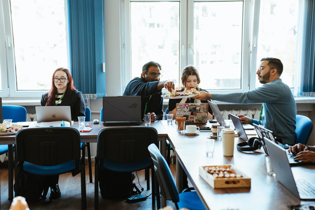 A group of people working at laptops in an office conference room as one person offers a plate of appetizers to another.