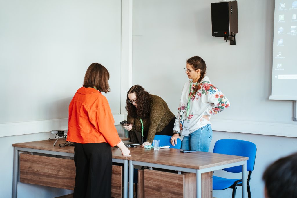 Three people stand around a desk in a classroom, with one person looking at a phone while the others observe.