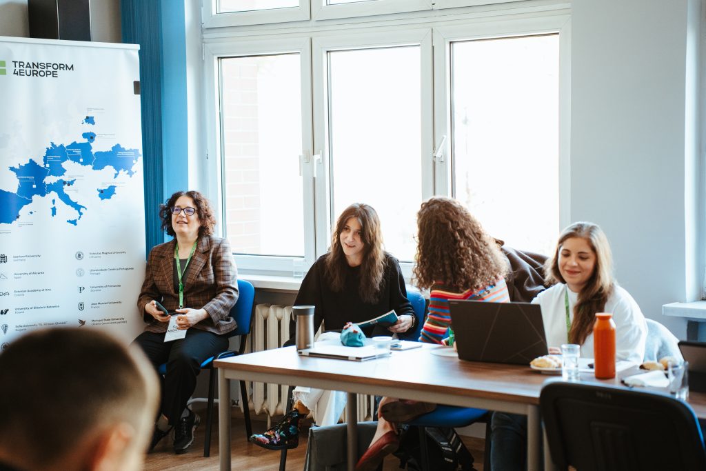 Four people sit around a table at a meeting, with a Transform4Europe banner featuring a map of European universities visible in the background.
