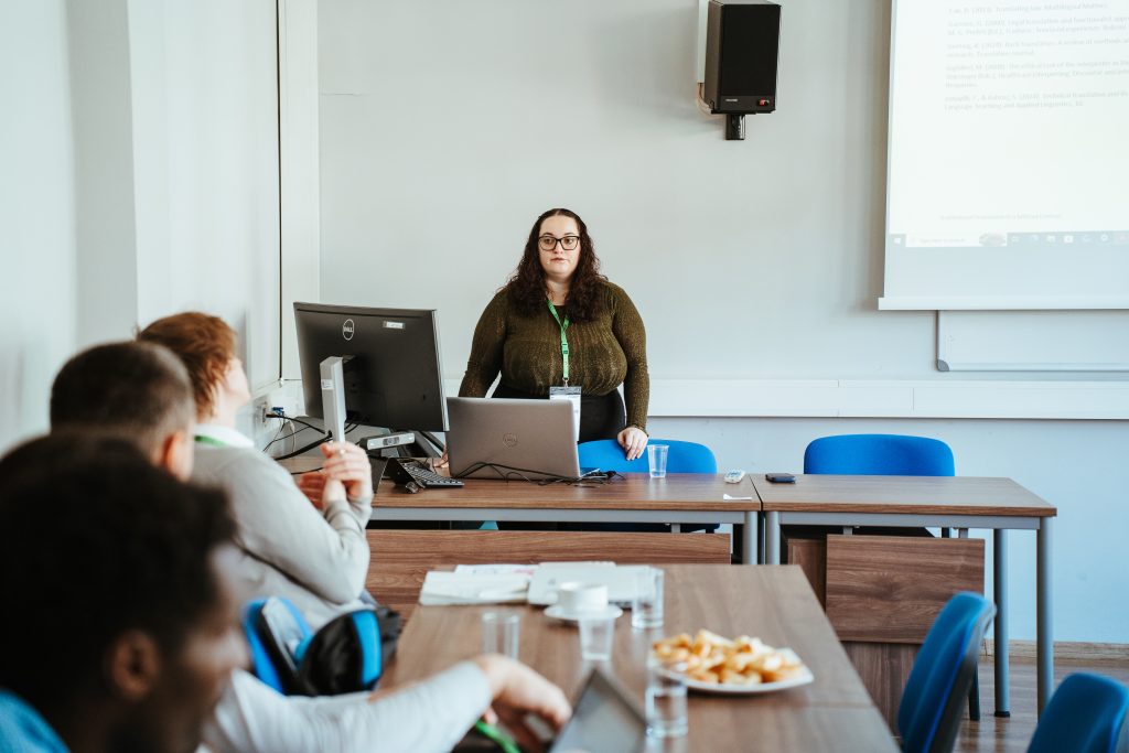 A woman wearing glasses and a lanyard stands behind a desk with a laptop and monitor, presenting to a group of students in a classroom.