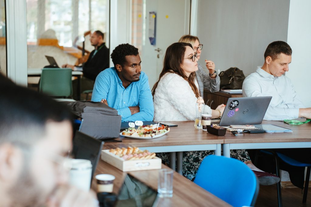 A group of people sit around a table in a workspace, listening to a meeting while snacks are laid out before them.