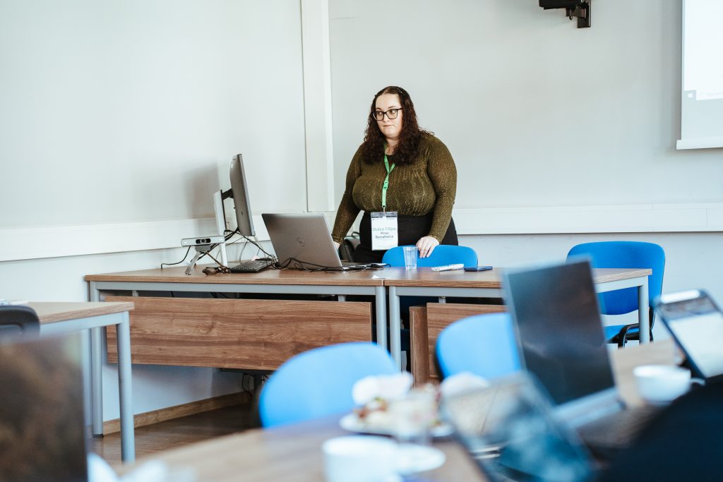 A woman wearing glasses and a conference badge stands behind a desk with a laptop in a classroom.