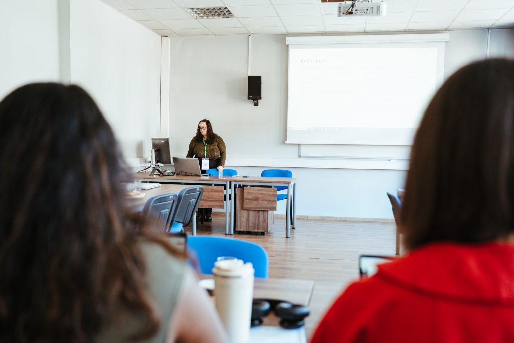 A woman presenting in front of a projector screen in a classroom, as viewed from behind two audience members.