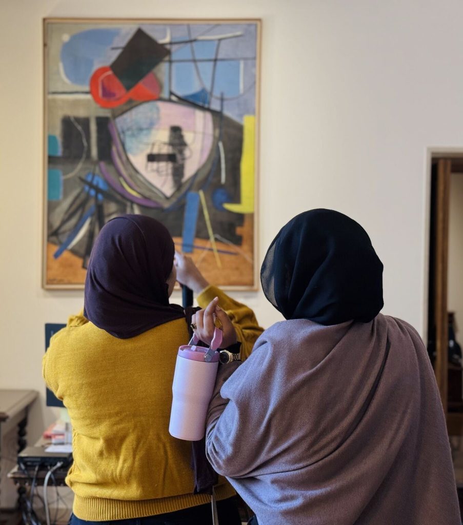 Two women wearing hijabs stand with their backs to the camera, looking at an abstract painting on a wall.