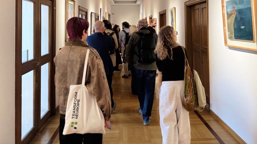 A group of people walk down a narrow hallway lined with framed paintings on the walls.