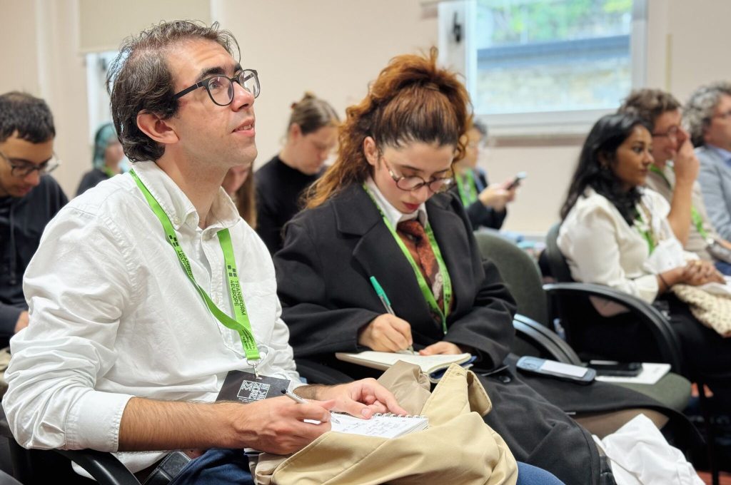A diverse group of people sitting in a classroom, with a man in the foreground taking notes while looking up toward the front of the room.