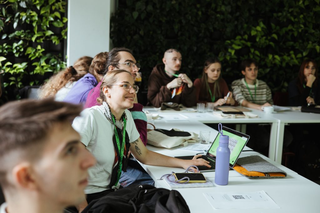 A group of people attending a meeting at white tables, with a woman in the foreground smiling while using a laptop.