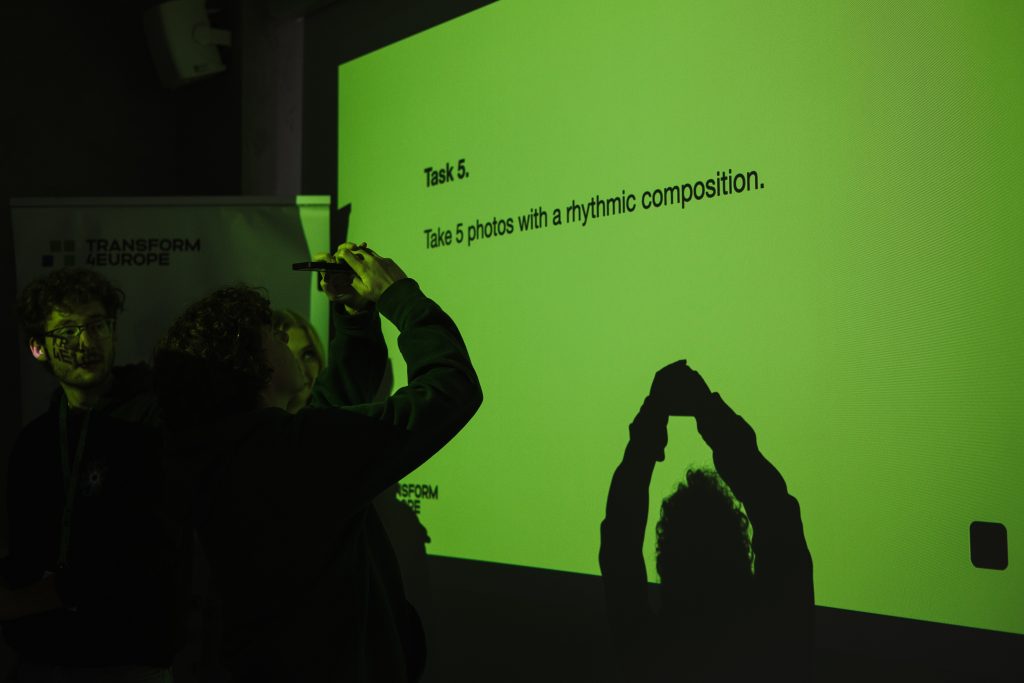 A person holds a phone up to photograph a bright green projection screen that reads, Task 5. Take 5 photos with a rhythmic composition.