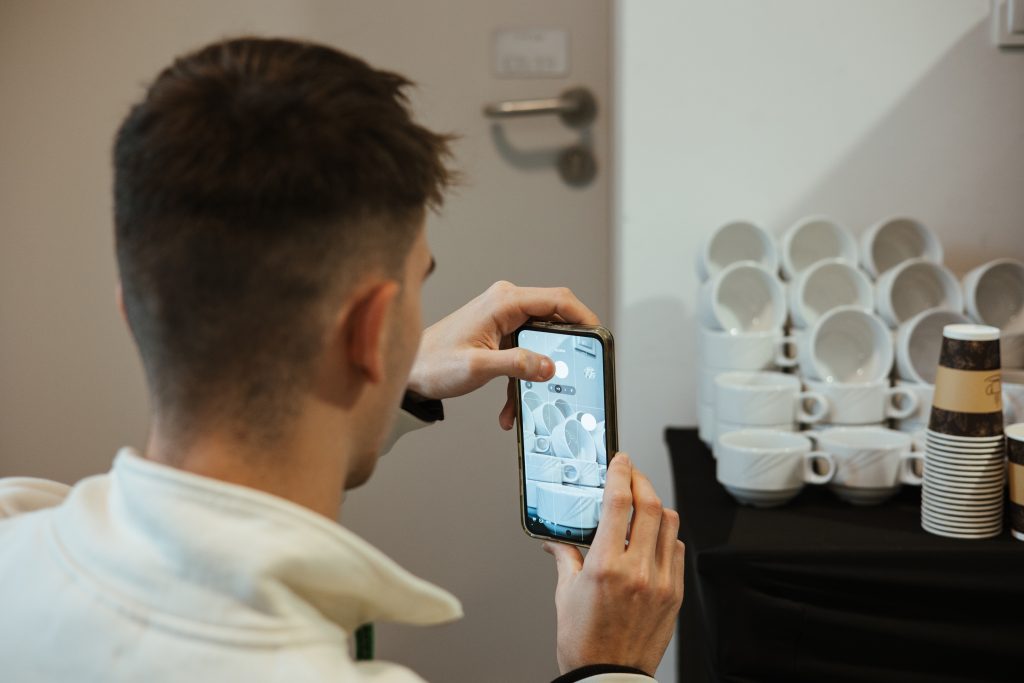A man uses a smartphone to take a photo of a stack of white coffee mugs on a table.