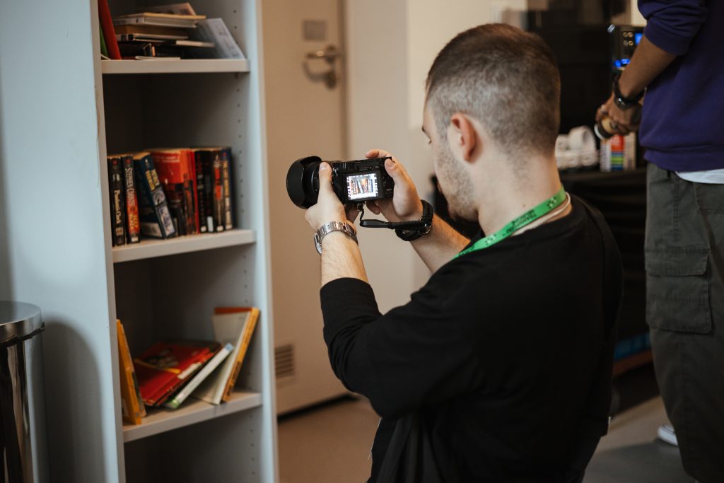 A person in a black long-sleeved shirt holds a digital camera to photograph books on a white shelf.