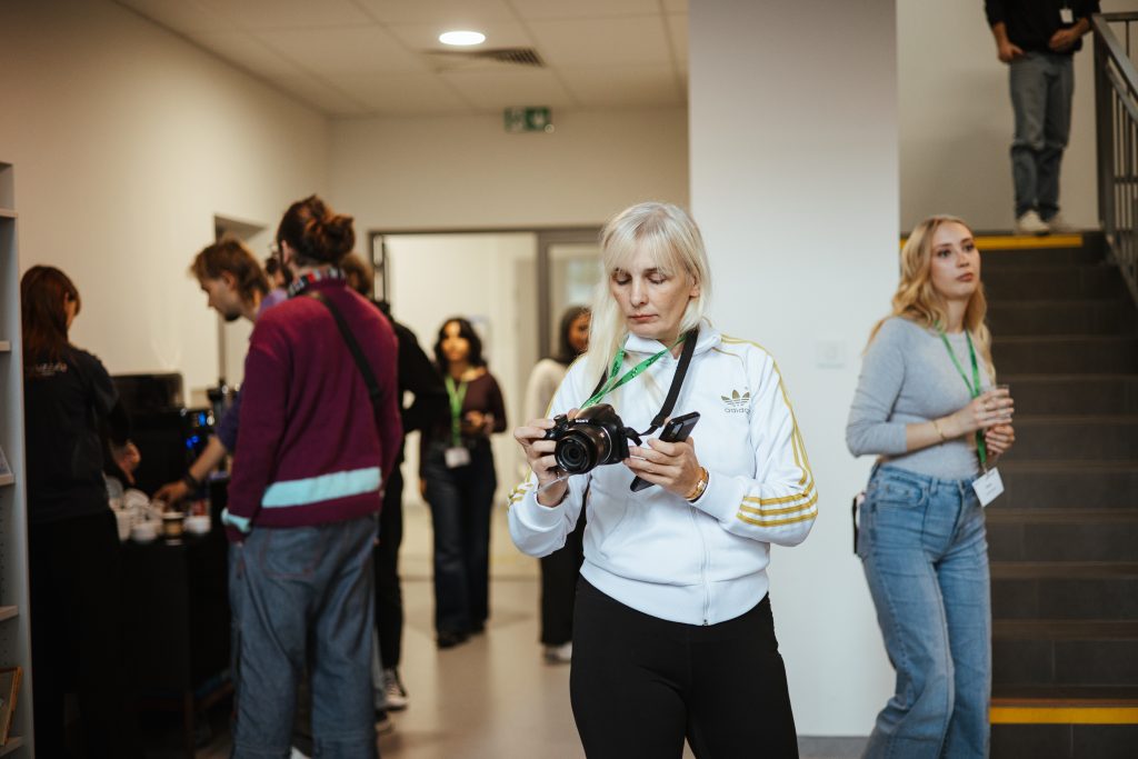 A woman in a white track jacket holds a digital camera while looking at a smartphone, with other people standing in an office hallway behind her.