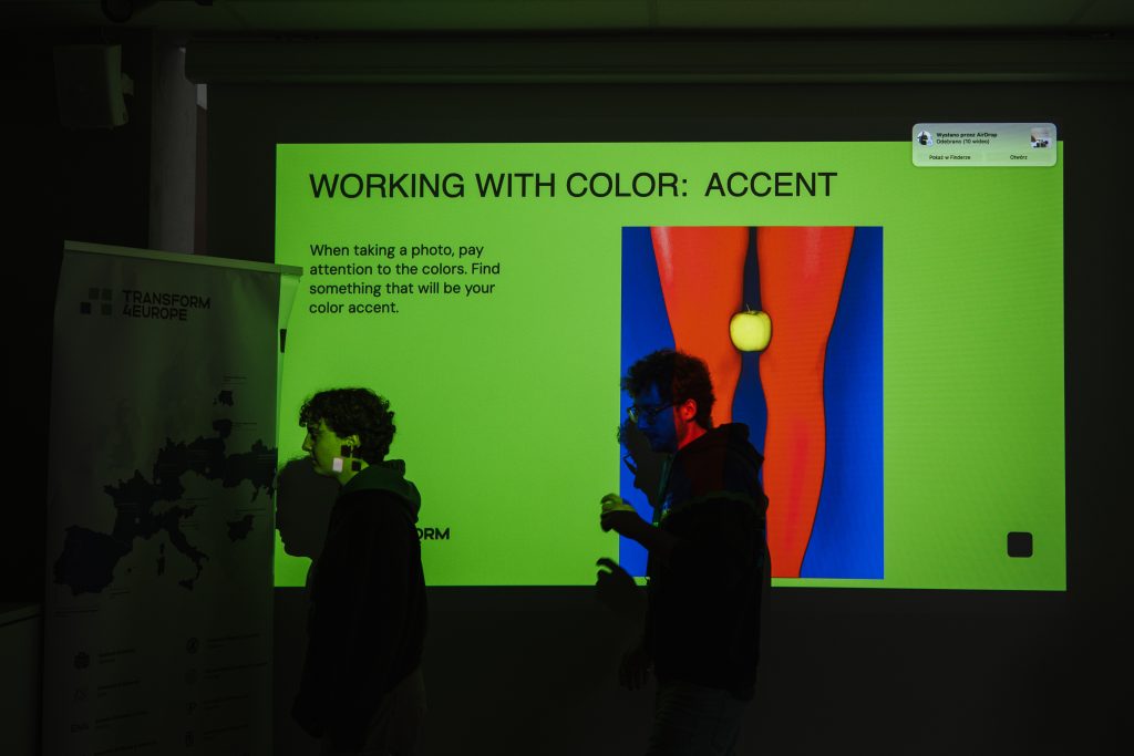 Two people walk in front of a projector screen displaying a presentation titled Working with Color: Accent, which features an image of a yellow apple positioned between two bright orange legs.