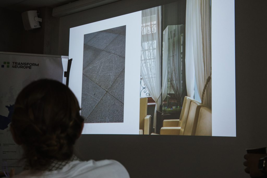 A person watches a presentation showing a side-by-side image of stone paving with a geometric line drawing and a brightly lit interior space with chairs and curtains.