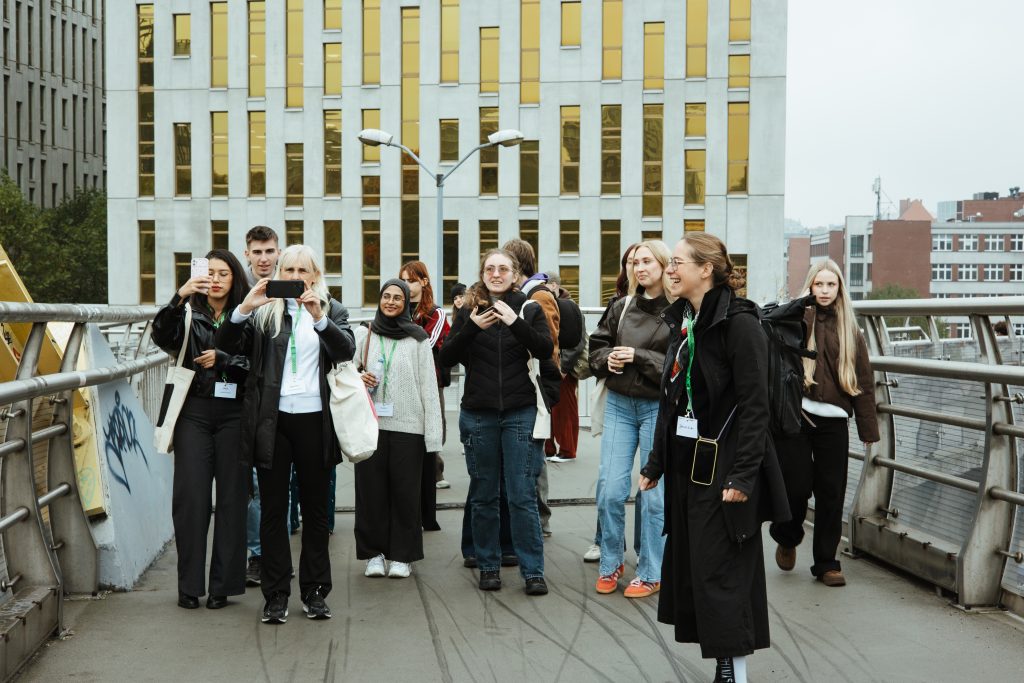 A diverse group of young people on a pedestrian bridge, with several individuals holding up their phones to take pictures of a building in the background.