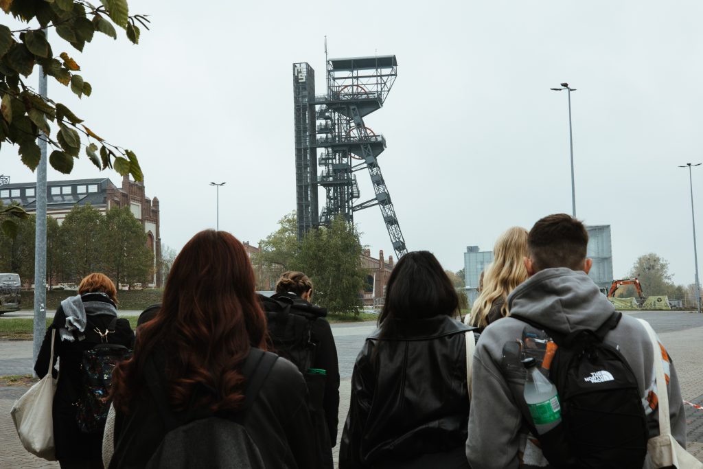 A group of people seen from behind walking toward a tall industrial mine headframe structure under an overcast sky.