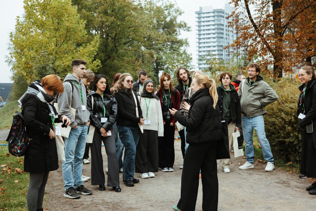 A woman speaks to a group of young adults standing in a circle outdoors in a park.