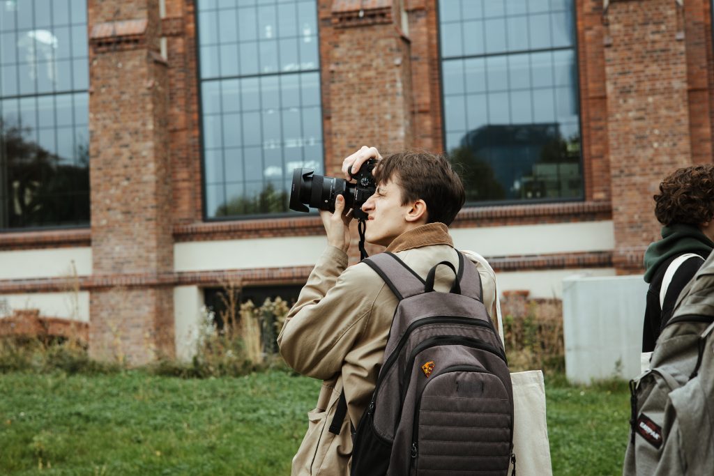 A person with a backpack holding a camera to their eye to take a picture in front of a brick building.