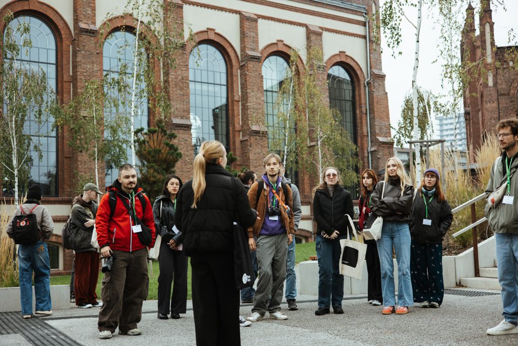 A group of people standing outside a brick building, listening to a woman speaking to them.