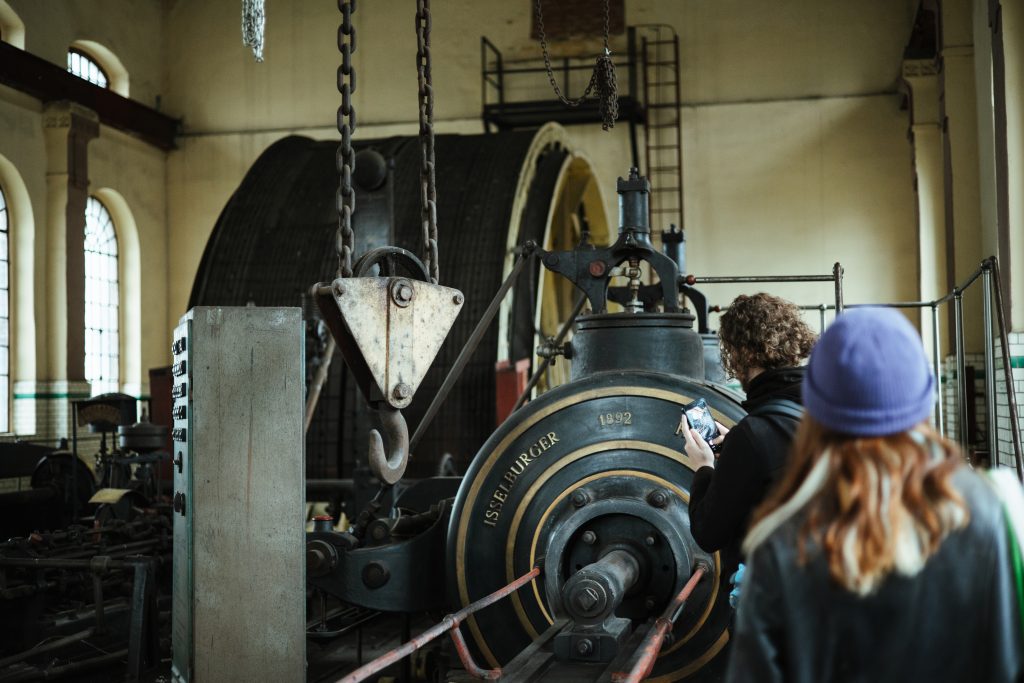 Two visitors take photos of large industrial machinery inside a workshop.