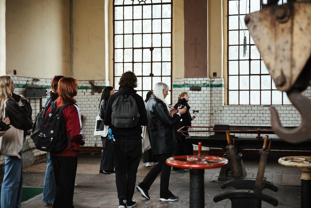 A group of people standing inside an industrial building with large windows, looking at machinery.
