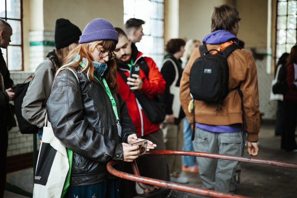 A person in a purple beanie and leather jacket looks at their phone while standing in a crowd of people.