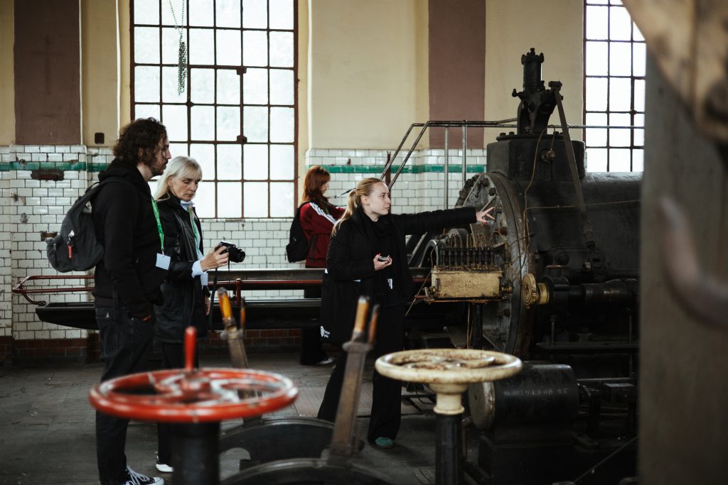 A tour guide points to a large piece of vintage industrial machinery while two visitors look on in an old factory building.