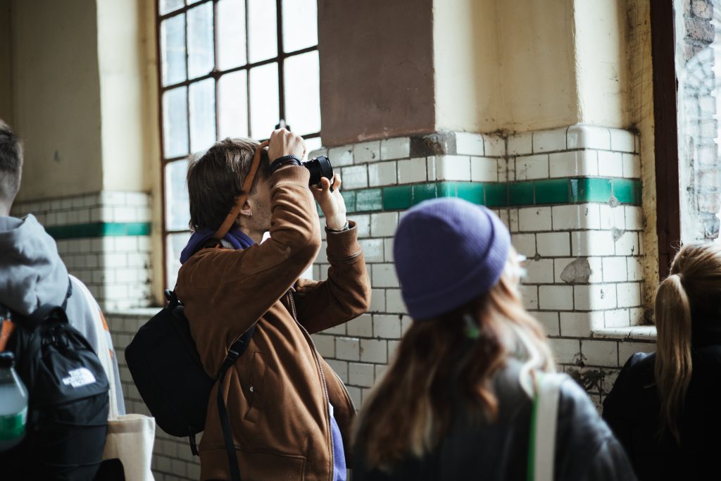 A person in a brown jacket uses a camera to take a photo of an industrial window, with other people standing nearby.