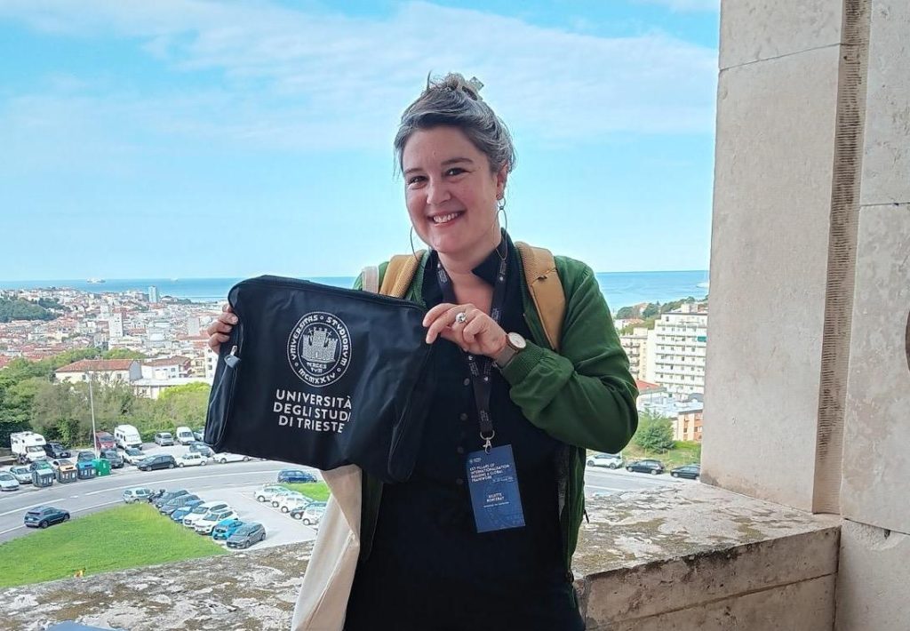 A woman smiling while holding a black bag featuring the University of Trieste logo, with an aerial city view in the background.