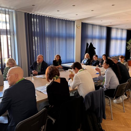 A group of people sit around a long conference table in a meeting room with blue blinds and sunlight streaming in. Papers, folders, and drinks are on the table, and everyone appears attentive and engaged in discussion.