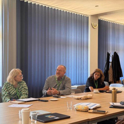 Five people sit around a conference table engaged in discussion. Papers, coffee cups, and water glasses are on the table. Coats hang on the wall behind them, and tall windows with blue vertical blinds let in daylight.