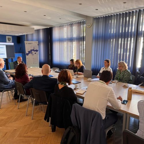 A group of people sit around a conference table in a meeting room with large windows, laptops, and documents. A screen at the front displays a presentation.