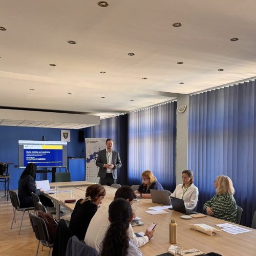 A man stands and presents to a group of people seated around a conference table in a bright meeting room. A screen displays a presentation in the background.