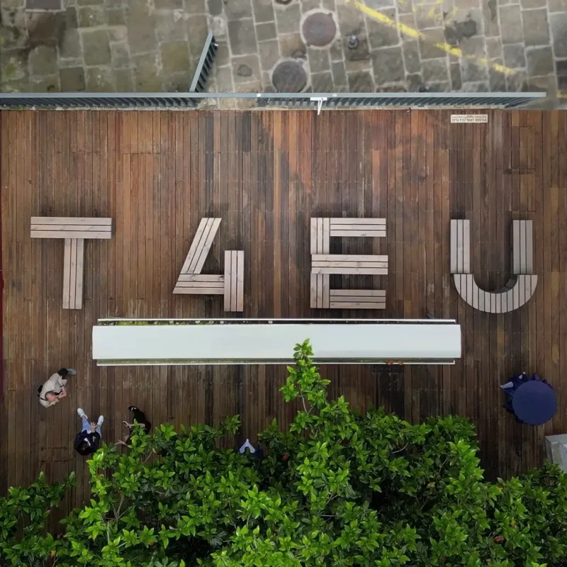 Aerial view of wooden benches arranged on a deck spelling out “T4EU,” with people walking nearby and green trees at the bottom of the image.