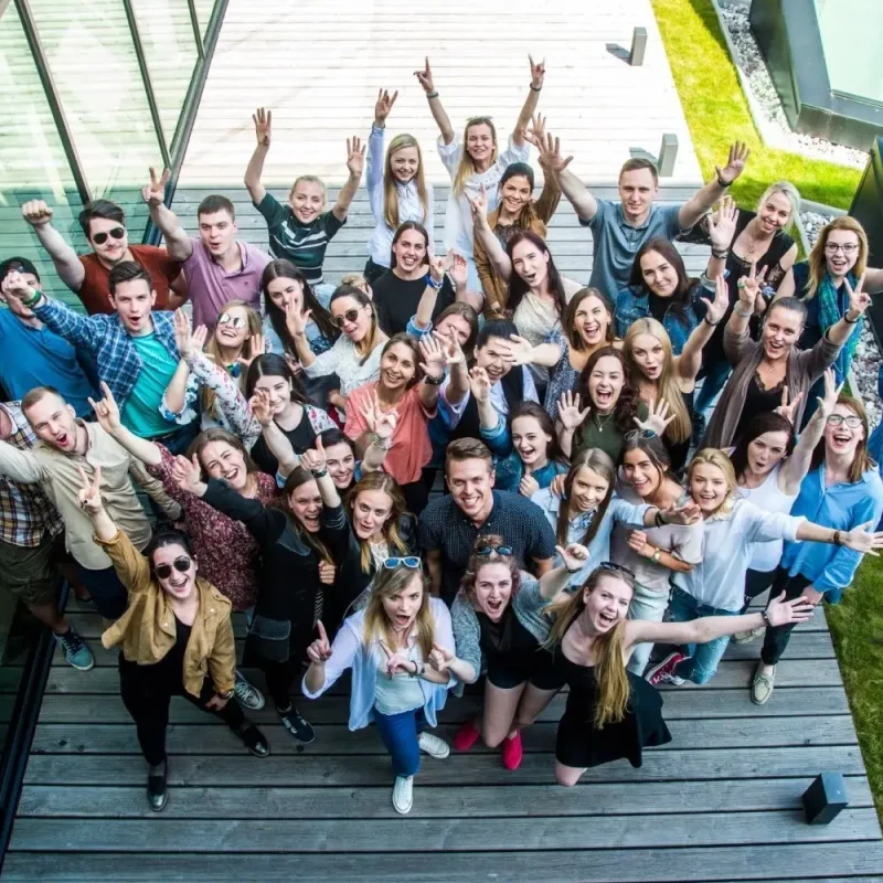 A large group of young adults stands close together outdoors on a wooden deck, smiling and raising their arms in celebration, looking up at the camera from above.